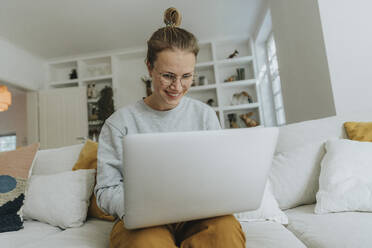 Lächelnde Frau arbeitet am Laptop, während sie zu Hause auf dem Sofa sitzt - MFF06199