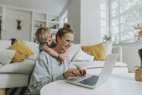 Woman working on laptop while boy hugging her from behind at home stock photo