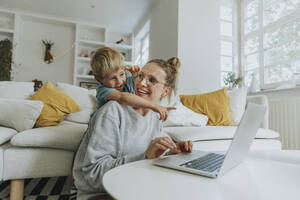 Junge zeigt auf Laptop, während er zu Hause hinter seiner Mutter steht - MFF06213