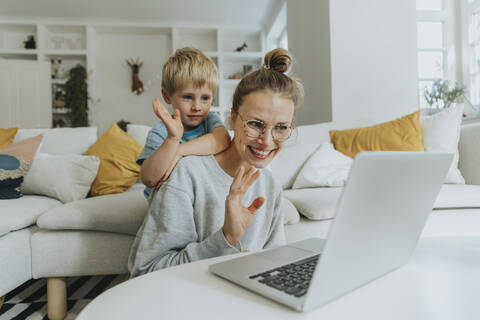 Mother and son waving hand to video call on laptop while sitting at home stock photo