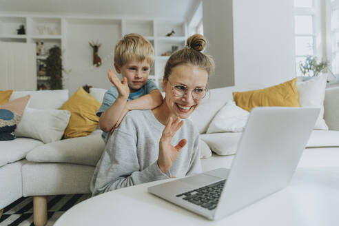 Mutter und Sohn winken mit der Hand zum Videoanruf auf dem Laptop, während sie zu Hause sitzen - MFF06215