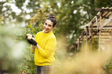 Smiling woman holding vegetable while standing at urban garden - SGF02686