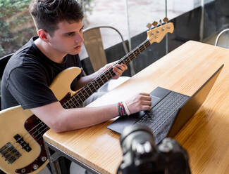 Von oben von konzentrierten jungen Mann sitzt am Tisch und beobachten Video-Tutorial auf dem Laptop beim Lernen, Musik auf der Bassgitarre zu spielen - ADSF15943