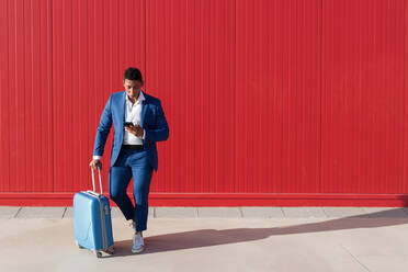 Ganzkörperaufnahme eines jungen afroamerikanischen Mannes in einem eleganten blauen Anzug mit Gepäck, der auf seinem Handy Nachrichten schreibt, während er an einer roten Wand auf der Straße steht - ADSF15944