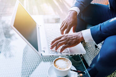 Seitenansicht eines anonymen schwarzen Geschäftsmannes im Anzug, der mit einer Tasse Kaffee am Tisch sitzt und auf einem Laptop tippt, während er in einem Café arbeitet - ADSF15945