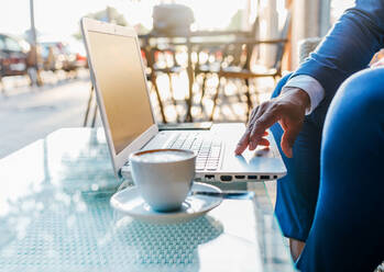 Seitenansicht eines anonymen schwarzen Geschäftsmannes im Anzug, der mit einer Tasse Kaffee am Tisch sitzt und auf einem Laptop tippt, während er in einem Café arbeitet - ADSF15946