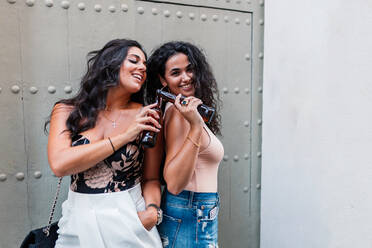 Positiv entspannte junge lateinamerikanische Freundinnen stoßen mit Bierflaschen an, während sie gemeinsam an einer grauen Wand chillen - ADSF15949