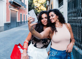 Fröhliche junge lateinamerikanische Freundinnen in stilvoller Kleidung stehen auf einer engen Straße und machen ein Selfie mit ihrem Smartphone, während sie einen Sommertag in der Stadt verbringen - ADSF15951