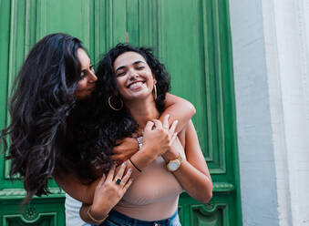 Positive entspannte junge lateinamerikanische Freundinnen umarmen sich auf der Straße vor einer grünen Tür - ADSF15952