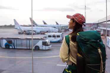 Rückenansicht einer jungen asiatischen Reisenden mit Rucksack, die in der Nähe des Fensters im Flughafenterminal steht und die Flugzeuge beobachtet, während sie auf ihren Flug in Sri Lanka wartet - ADSF15957