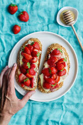 Draufsicht auf eine nicht erkennbare Frau, die einen leckeren süßen Brottoast mit frischen gehackten Erdbeeren und Honig auf einem Teller auf einem blauen Tischtuch serviert - ADSF15979