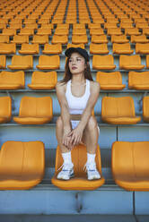 Confident Asian athletic woman in sportswear sitting on stadium seat while resting after workout and looking away - ADSF15983