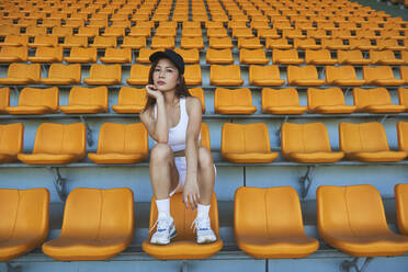 Confident Asian athletic woman in sportswear sitting on stadium seat while resting after workout and looking at camera - ADSF16077