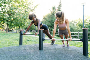 Ganzkörper positive junge multirassische Freundinnen in Sportkleidung machen Push-up-Übung auf Metallstangen während Fitness-Workout im Sommer Park - ADSF16091