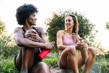 Niedriger Winkel von lächelnden jungen multiethnischen Freundinnen in Sportkleidung, die Wasser trinken und sich unterhalten, während sie auf einem Stein sitzen und sich nach dem Training in der Natur ausruhen - ADSF16092