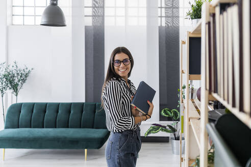 Glückliche schöne junge Design-Profi hält Buch, während stehend von Rack auf kreative Büro - GIOF08975