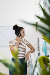 Smiling young female architect talking on mobile phone while standing by table at creative office - GIOF08989