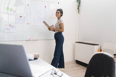 Smiling beautiful young female architect holding clipboard while standing by whiteboard in creative workplace - GIOF08991