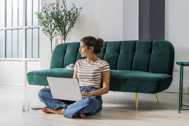 Nachdenkliche Architektin, die im Schneidersitz auf dem Boden sitzt und einen Laptop auf dem Sofa im Büro benutzt - GIOF09015