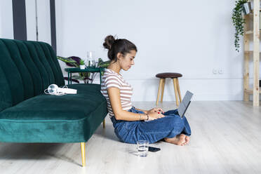 Selbstbewusste Architektin, die einen Laptop benutzt und im Schneidersitz auf dem Boden vor dem Sofa im Büro sitzt - GIOF09018