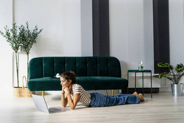 Smiling businesswoman looking at laptop while lying on floor by sofa in creative workplace - GIOF09020