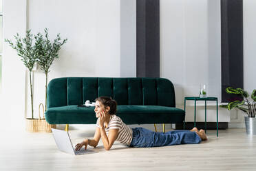 Thoughtful female architect looking away while lying with laptop on floor by sofa in creative workplace - GIOF09021