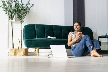 Thoughtful female design professional holding coffee cup while looking away and sitting by laptop against sofa at office - GIOF09022