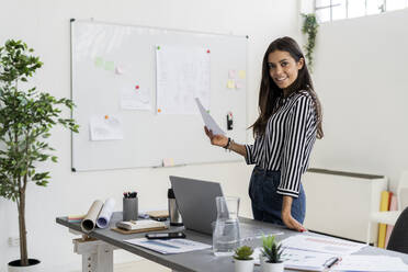 Lächelnde schöne Geschäftsfrau, die einen Plan hält, während sie am Schreibtisch in einem kreativen Büro steht - GIOF09027