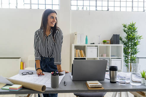 Lächelnde Geschäftsfrau, die wegschaut, während sie am Schreibtisch in einem kreativen Büro steht - GIOF09030