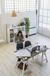 Female design professional photographing floor plan at desk in creative workplace - GIOF09035