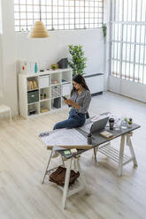 Young businesswoman using smart phone while sitting by blueprint at desk in creative workplace - GIOF09036