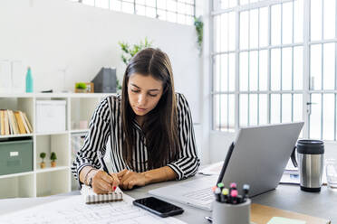 Beautiful businesswoman writing in diary while sitting at desk in office - GIOF09039
