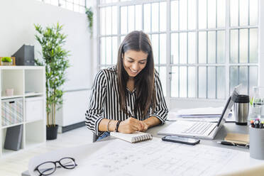 Lächelnde junge Architektin, die am Schreibtisch im Büro sitzt und in ihr Tagebuch schreibt - GIOF09040