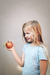 Cute blond girl holding fresh organic red apple against wall - IPF00553