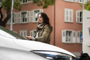 Selbstbewusste schöne Frau, die ein Smartphone benutzt, um ein Elektroauto an einer Ladestation zu mieten - SGF02696