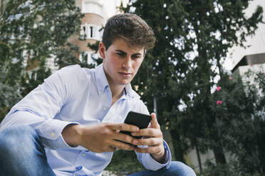 Hübscher junger Mann benutzt sein Smartphone, während er in der Stadt an einem Baum sitzt - ABZF03339