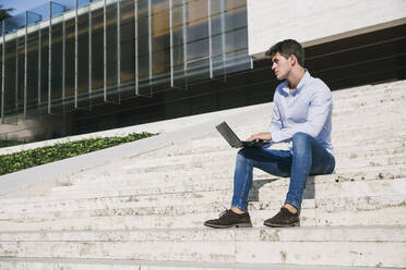 Nachdenklicher Mann schaut weg, während er mit seinem Laptop auf einer Treppe in der Stadt sitzt, während eines sonnigen Tages - ABZF03340