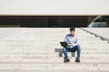 Selbstbewusster junger Mann, der einen Laptop benutzt, während er an einem sonnigen Tag auf einer Treppe sitzt - ABZF03341