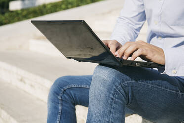 Man typing on laptop while sitting outdoors during sunny day - ABZF03343