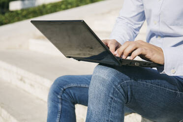 Mann tippt auf einem Laptop, während er an einem sonnigen Tag im Freien sitzt - ABZF03343