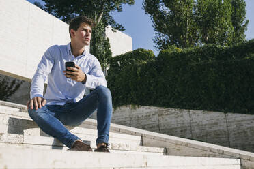 Nachdenklich gut aussehend junger Mann sitzt mit Smartphone auf Stufen gegen Bäume in der Stadt während sonnigen Tag - ABZF03347