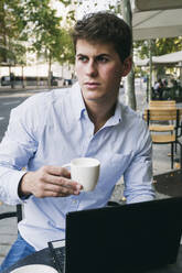 Nachdenklicher junger Mann hält eine Kaffeetasse, während er mit seinem Laptop in einem Straßencafé sitzt - ABZF03349