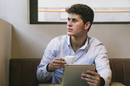 Gut aussehender junger Mann mit Kaffeetasse und digitalem Tablet, der in einem Café sitzt - ABZF03350