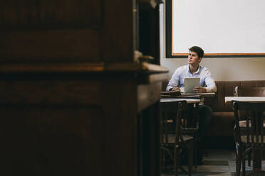Nachdenklicher Mann, der eine Kaffeetasse und ein digitales Tablet hält, während er in einem Café sitzt - ABZF03351