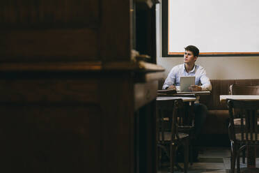 Thoughtful man holding coffee cup and digital tablet while sitting at cafe - ABZF03351