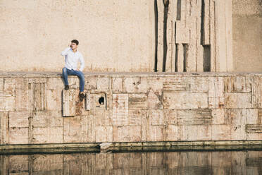 Gut aussehender junger Mann, der mit seinem Mobiltelefon spricht, während er an einem sonnigen Tag auf einer Mauer über einem Kanal sitzt - ABZF03353