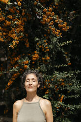 Reife Frau mit geschlossenen Augen vor einem Orangenbaum an einem sonnigen Tag stehend - TCEF01172