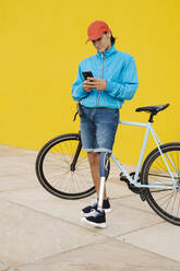 Man using phone while standing by bicycle against yellow wall - JCZF00260