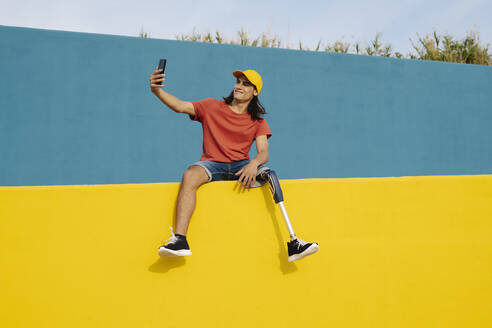 Junger Mann, der ein Selfie macht, während er auf einer mehrfarbigen Wand sitzt - JCZF00281