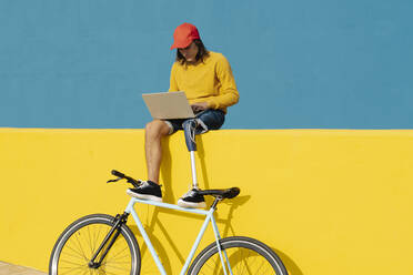 Man with artificial limb and foot working on laptop while sitting on multi colored wall - JCZF00288